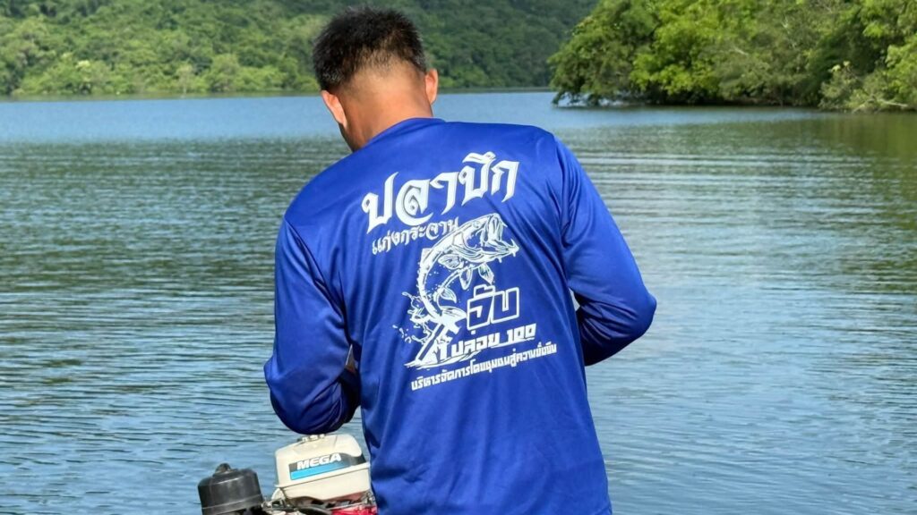 ด้านหลังเสื้อชาวประมงพื้นบ้านเขื่อนแก่งกระจาน สกรีนข้อความ "ปลาบึก เขื่อนแก่งกระจาน จับ 1 ปล่อย 100 บริหารจัดการโดยชุมชนสู่ความยั่งยืน" ขณะยืนอยู่บนเรือหางยาวกลางอ่างเก็บน้ำ
